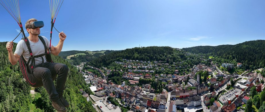 Triberg als Drohnenaufnahme und ein Gleitschirmflieger mit einer VR-Brille ist zu sehen.